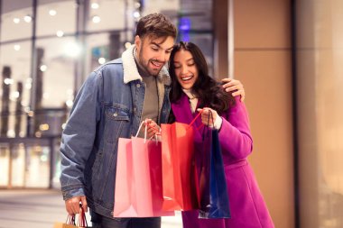 Genç çift sevgililer günü alışverişlerini sevinçle sergiliyor. Kış gecesi alışveriş merkezinin önünde birlikte duran çantaların içine bakıyorlar. Kutlama satışları ve indirimler konsepti