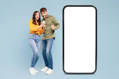 Neşeli erkek ve kadın akıllı telefondan keyif alıyorlar, büyük boy bir telefonun yanında dikiliyorlar, mavi bir arkaplanda duruyorlar, dijital reklam için ideal.