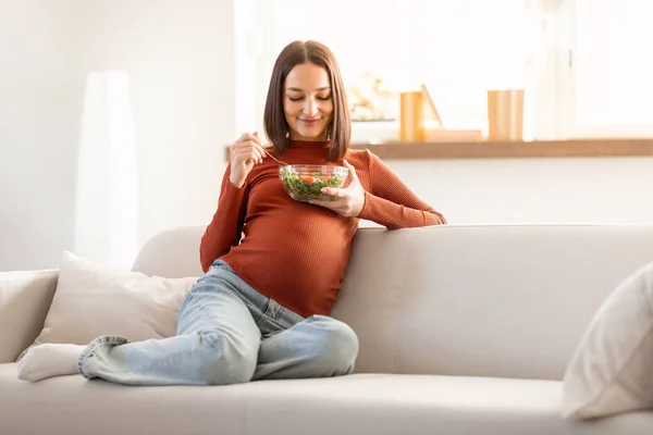 Healthy Pregnancy Nutrition. Glad Pregnant Woman Eating Fresh Veggie Salad While Sitting On Sofa Indoor, At Modern Home Interior. Holding Bowl With Tasty Organic Food, Enjoying Expectation Menu