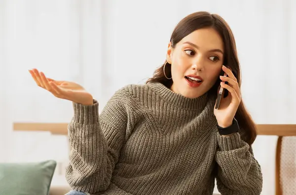 Closeup of talkative young brunette woman talking on phone and gesturing while sitting on couch in cozy living room interior, sharing rumors with her girlfriend, copy space