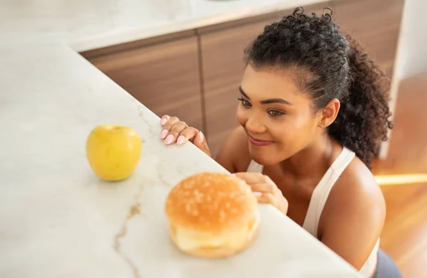 Food choice. Hungry fit lady looking at unhealthy burger and healthy apple, choosing her meal near kitchen table indoors. Sporty woman chooses between balanced and unbalanced nutrition