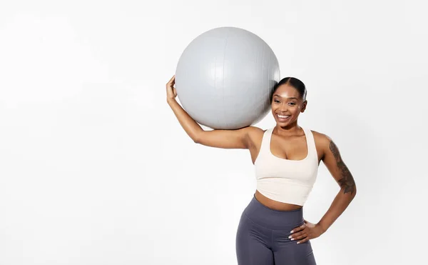 健身健身锻炼 身穿运动服的黑人女运动员站在那里拿着稳定球 对着镜头微笑 展示她的力量和平衡 摆出一副白色工作室的样子 空白的文字空间 — 图库照片