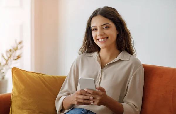 Beautiful Young Smiling Woman With Smartphone In Hands Relaxing On Couch At Home, Happy European Female Using Mobile Phone For Communication Or Online Shopping, Looking At Camera, Copy Space