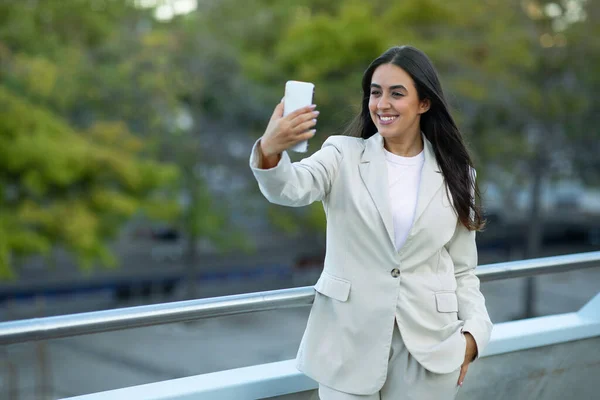 Positive Arabic businesswoman making video call on smartphone gadget, standing outside in modern urban area, managing remote communication for her job. Free space for text