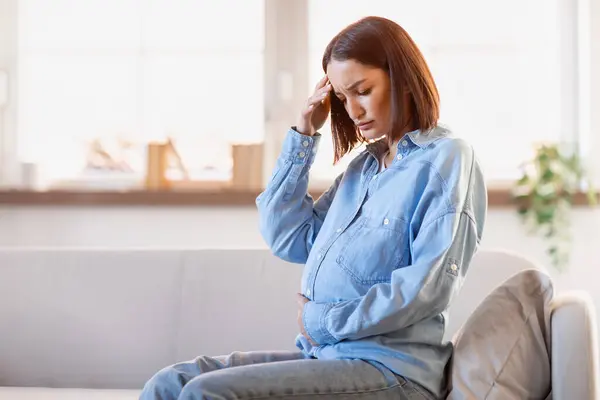 Ill young pregnant woman touches forehead in discomfort, feeling unwell suffering from headache and fever symptoms sitting on couch at home. Challenges of pregnancy health issues