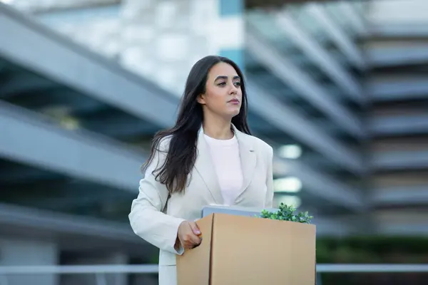Sad Hispanic business lady carrying cardboard box with her belongings, leaving office building after layoff, reflecting economic challenges of corporate dismissal and unemployment