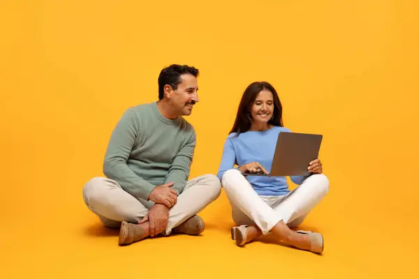 A man and woman sit cross-legged, woman use laptop and both looking at screen with interest, collaboration and shared focus on orange background. Watch video, recommendation