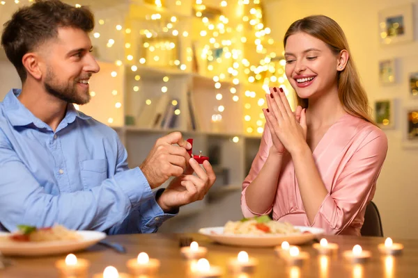 Joyful man in blue shirt presents engagement ring to excited woman in pink, candles and twinkling lights creating romantic atmosphere at home
