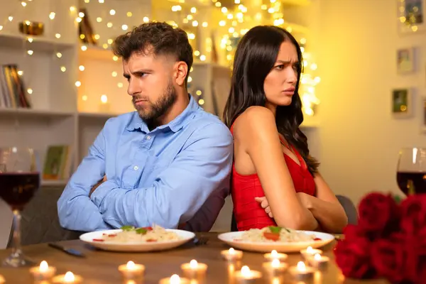 Unhappy couple in disagreement, sitting with arms crossed and back-to-back over dinner, amidst romantic setting turned sour