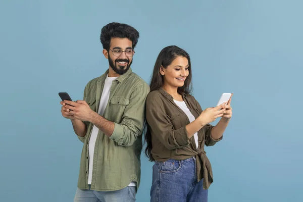 Smiling young indian man with smartphone in his hands spying on girlfriend using cell phone, eastern guy sending his wife funny reels, isolated on blue studio background