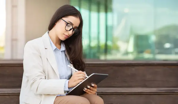 Focused caucasian millennial woman in glasses, sits outside, deeply engaged in writing on a digital tablet, suggesting productivity and technology use. Tech-savvy professionalism and focus