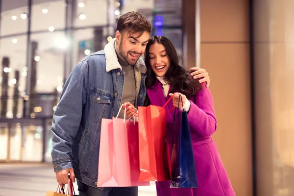 这对年轻夫妇兴高采烈地展示了他们的情人节购物 在冬夜 他们一起站在一个城市购物中心外面的袋子里偷窥着 节日销售和折扣概念 — 图库照片