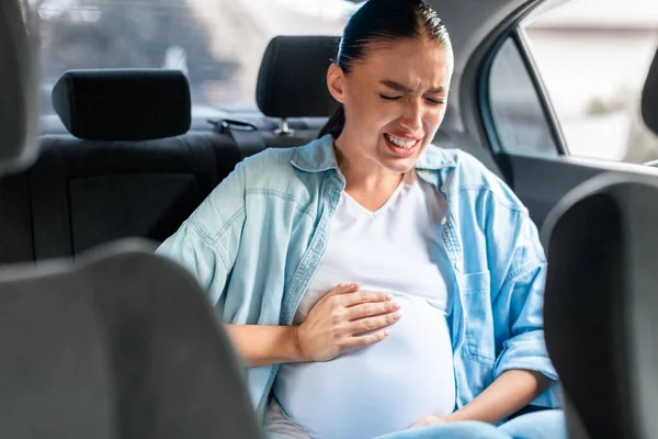 Pregnant lady in car suffering from labor pains clutching her abdomen while sitting in back seat of auto. Healthcare and transportation problems during pregnancy. Selective focus