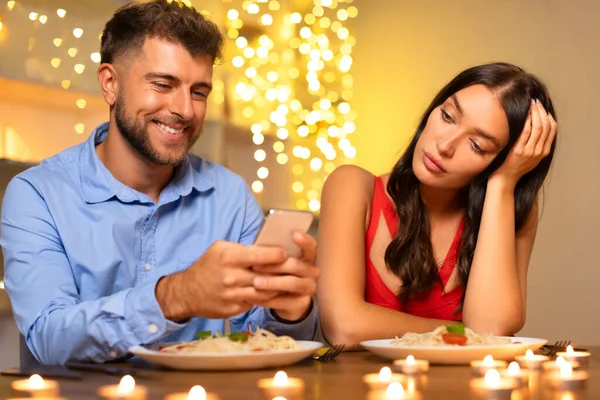 Cheerful man engrossed in smartphone, disinterested woman with hand on cheek during romantic candlelit dinner, hint of frustration in ambiance