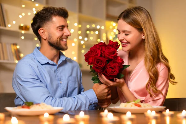 Beaming with joy, man presents a lush bouquet of red roses to a woman, adding a touch of romance to their candlelit dinner setting