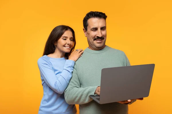 Smiling european man in sweater and woman look at laptop screen together, lady resting hand on his shoulder, both displaying expressions of interest and pleasure on orange background