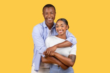 Happy, attractive black couple in close embrace, sharing genuine smiles with the camera, set against monochromatic yellow background