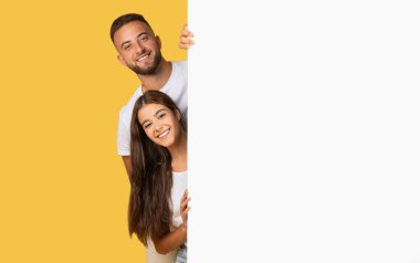 A young european man and woman peek around a blank white sign with joyful expressions, set against a warm yellow studio background, inviting engagement. Attention, good news, sale