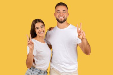 Radiant young european couple in white shirts showing peace signs with bright smiles, projecting a vibe of harmony and positivity gesture against a yellow studio backdrop