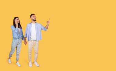 Excited man pointing finger up aside at copy space, walking holding hands with surprised woman, demonstrating free place for advert, yellow studio background, banner