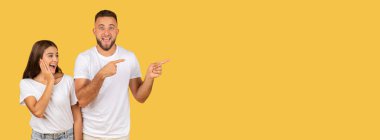 Excited happy young caucasian man and woman in white t-shirts point to the side with wide smiles, sharing a fun moment on a solid yellow studio background, panorama. Attention, good news, sale