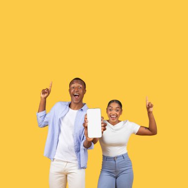 Joyful black couple pointing upwards and presenting smartphone screen with blank space for content, embodying excitement and surprise, against yellow background, mockup