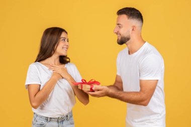 A joyous young caucasian woman looks at a smiling man who is giving her a small gift box with a red ribbon, representing a gesture of affection and surprise on a yellow background