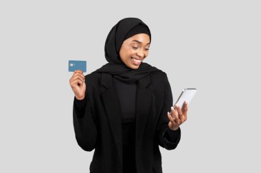 Smiling young african american lady in hijab use smartphone and credit card, enjoy online shopping, isolated on gray studio background. Banking app, cashback and finance money check