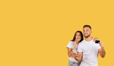 Intrigued young caucasian woman holding a smartphone and looking up with curiosity while a cheerful man beside her shows a credit card, both set against a yellow background