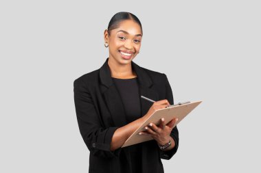 Positive pretty young african american lady secretary, manager in formal suit make notes at clipboard, isolated on gray studio background. Business, work and study, ad and offer