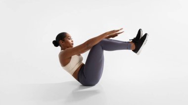 Focused African American woman exercising in boat pose outstretching arms, flexing abs muscles, showcasing fitness and balance in white studio, sitting on floor. Panorama, side view