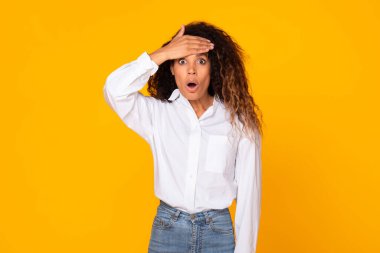 Shocked young black lady touching forehead and looking at camera with overwhelmed expression, having fever or stressing about unexpected news and problems, over yellow studio background