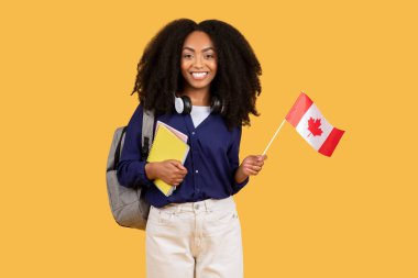 Cheerful black lady student, carrying backpack and copybooks, holds Canadian flag on yellow background, representing her pursuit of foreign language education and cultural understanding