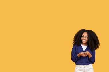 Cheerful black lady in casual using smartphone, looking at copy space for mobile application advertisement and smiling, yellow studio background, banner