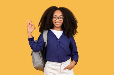 Happy black female student with backpack in glasses shows ok sign with hand isolated on yellow studio background. Recommendation of good study and knowledge, education