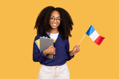 Excited african american lady student, with tablet and copybooks, holds flag of France and smiling on yellow background, enjoying foreign language education