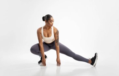 Fit black lady making extended leg squat pose stretching legs, exercising against white background. Full length studio shot of athlete woman having workout for muscles flexibility