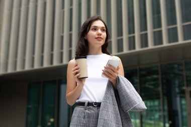 Young businesswoman browsing on smartphone, enjoying coffee on city street outdoor, holding her jacket and looking aside, embodying successful urban professional taking brief pause from work