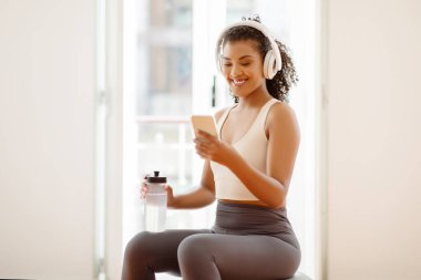 Smiling fit young woman in sportswear and headphones checks phone while holds water bottle, taking break during home workout, sitting on fitness ball indoors, browsing sport applications