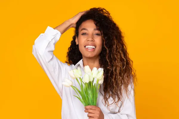 Happy young black lady holds bouquet of white tulips, smiling to camera enjoying spring holiday, isolated on yellow studio background. Gift to women day, congratulation on anniversary or date