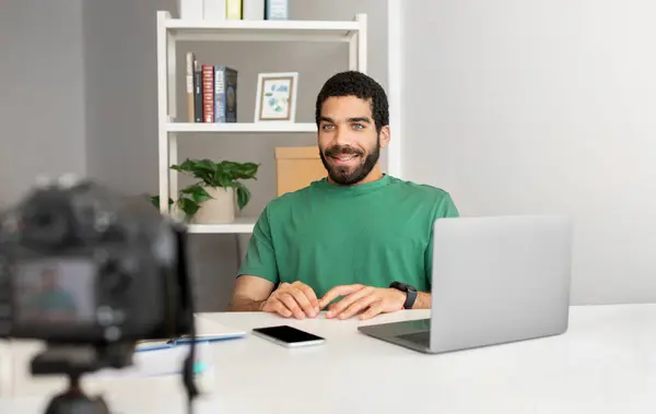 Smiling man in green shirt sitting at desk with a laptop, smartwatch, and smartphone, preparing for video recording session with a camera set up in front of him, in a well-organized office space