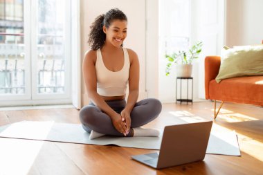 Online yoga class. Happy fit woman looking at laptop while sitting on fitness mat, ready for distance gym training and workout in modern living room at home. Sporty morning on weekend