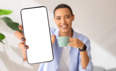 Focus on a smartphone screen held by a smiling woman, with a blurred background showing her holding a coffee cup, smartwatch on wrist, in a light, plant-filled room, blurred
