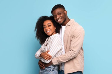 Cheerful young black couple in close embrace, sharing bright smiles and moment of affection, set against tranquil light blue background