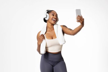 Happy african american lady in sportswear and wireless headphones making video call or selfie shot on smartphone, standing isolated on white studio background. Sport, workout and technology