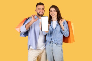 Happy family with many packages showing cellphone with empty screen, isolated on yellow studio background. Online banking app, shopping with technology, sale for shopaholic