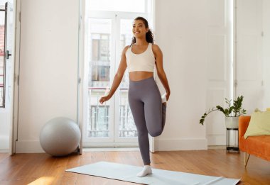 Young sporty woman stretching leg making heel to butt exercise in modern living room at home, starts her morning workout with warmup training. Healthy fitness. Full length shot