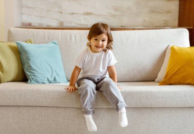 Cheerful little girl lounging on sofa at home, grinning brightly at camera, cute preschooler female child relaxing on on comfortable couch in living room, capturing the essence of youthful exuberance