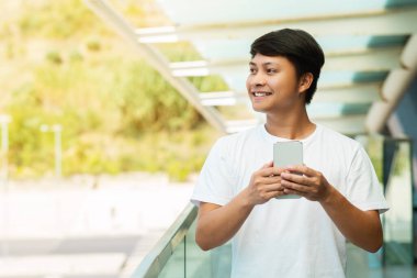 Positive young asian guy wearing white t-shirt with smartphone in his hands posing on the street, looking at copy space for advertisement, walking by city center, checking online adviser