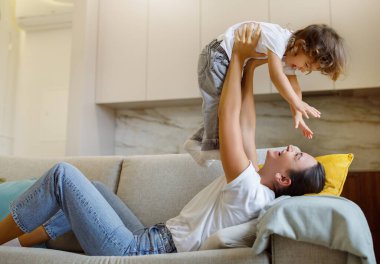 Mother and child playing airplane game on sofa, mom lifting her giggling daughter high, their faces lit with joy, mommy and female kid having fun together while relaxing on cozy sofa in living room
